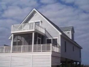 Gray beach house with white railings and porch, set against a cloudy blue sky.