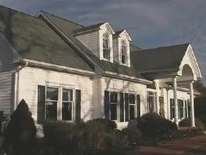 White house with dark shutters, green roof, and small dormers under a cloudy sky.