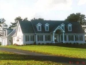 A light-colored house with a dark roof and a long driveway, on a grassy hill.