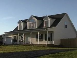 White house with a porch and three dormer windows, on a grassy lawn with a gravel driveway.
