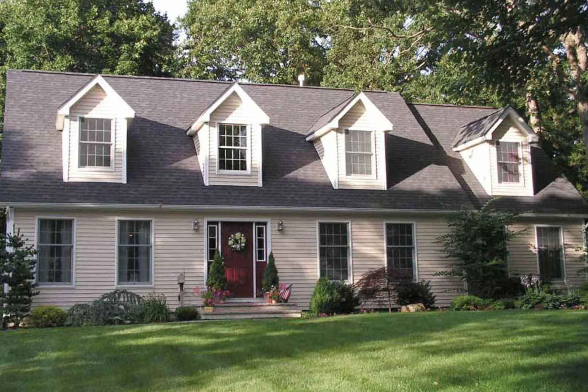 Tan house with black roof and four dormers. Front yard with green lawn and bushes.