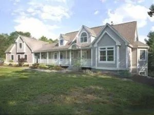 Gray house with porch and dormers, set on a grassy lawn under a partly cloudy sky.