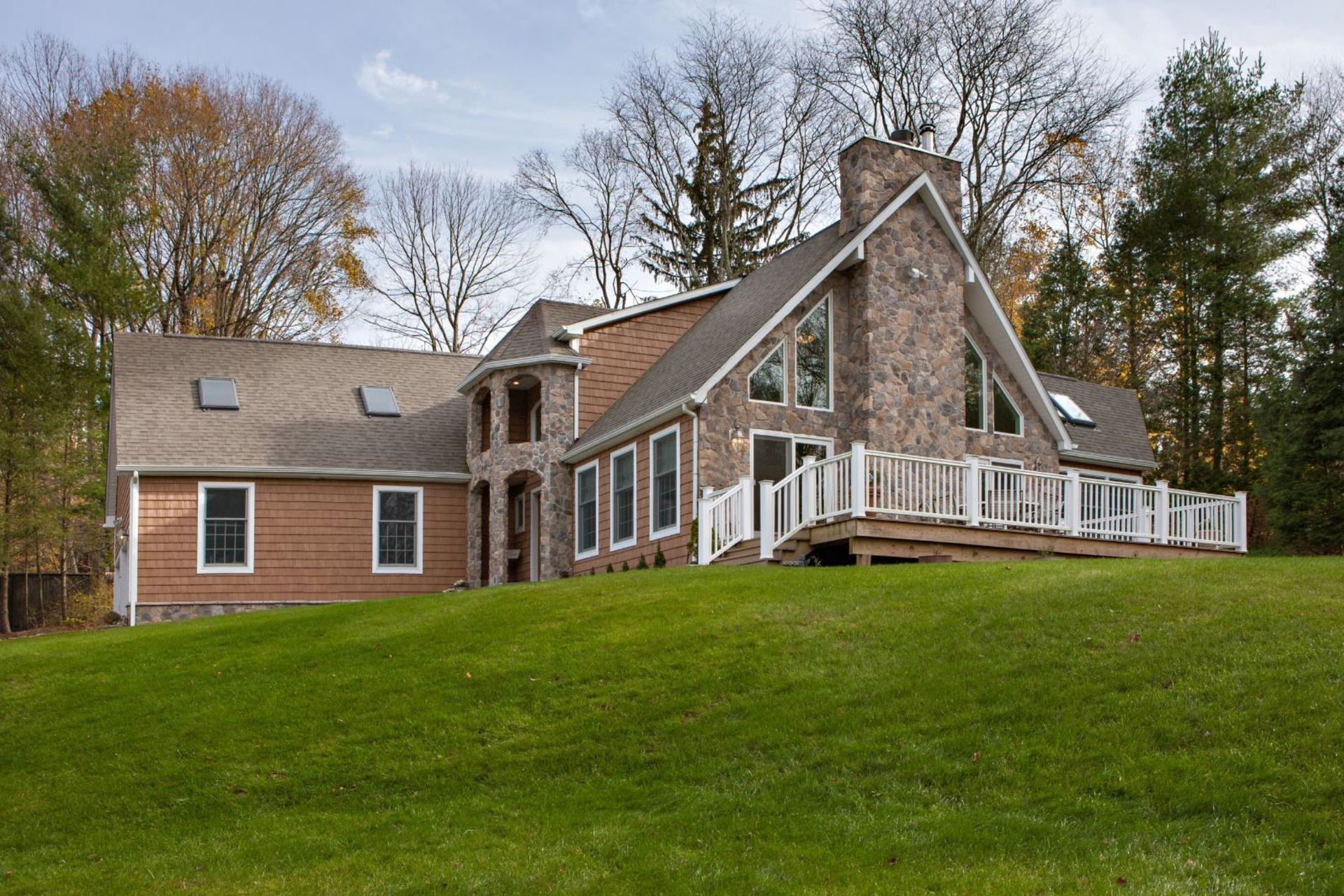 House with stone facade, brown siding, and deck on a green lawn, surrounded by trees.