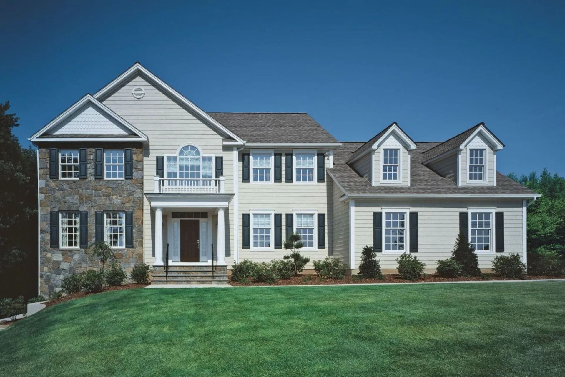 Two-story house with beige siding, stone facade, black shutters, green lawn, blue sky.