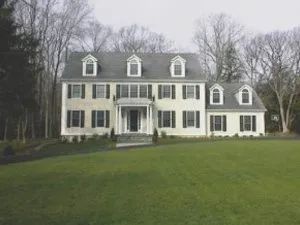 Two-story cream-colored house with black shutters, dormers, and a green lawn. Trees in the background.