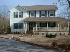 Two-story white house with front porch and green shutters on windows. Brown roof. Driveway in foreground.