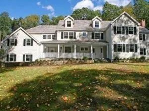Large white house with black shutters, green lawn, trees, and blue sky.