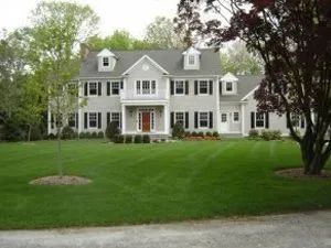 Two-story gray house with black shutters, green lawn, trees, and blue sky.
