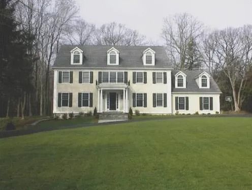 Two-story colonial house with tan siding, black shutters, and dormer windows, set on a green lawn.