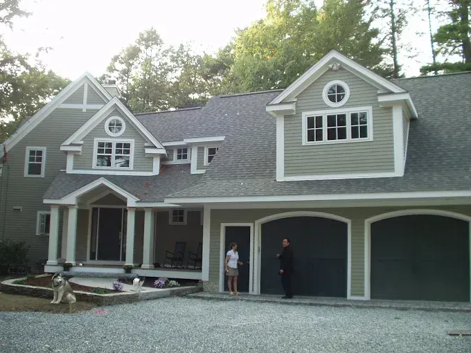 Two-story green house with a garage, two people stand near a door; a dog sits on a nearby wall.