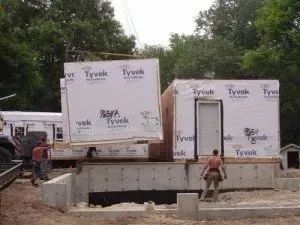 Workers lifting a modular home section with a crane, placing it on a concrete foundation; construction site.