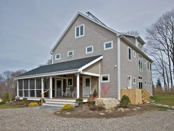 Two-story gray house with screened porch, wooden deck, and gravel driveway on a cloudy day.