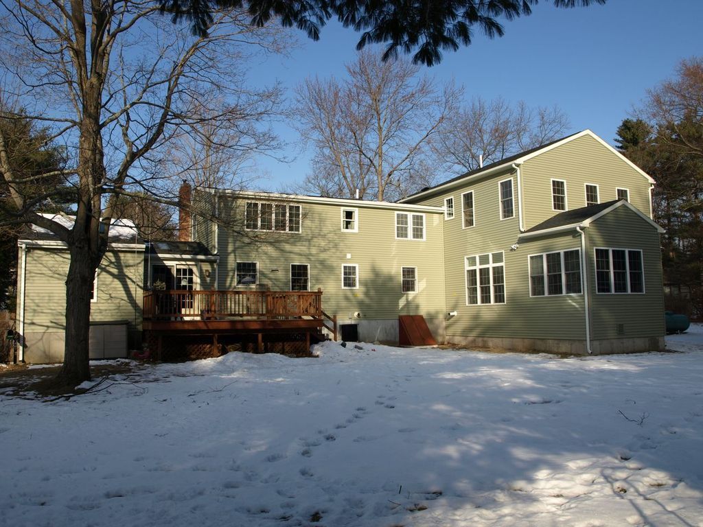Back of a two-story green house with a wooden deck, trees, and snow-covered ground.