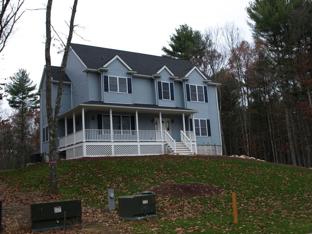 Two-story blue house with white porch and railing, on a grassy hill, surrounded by trees.