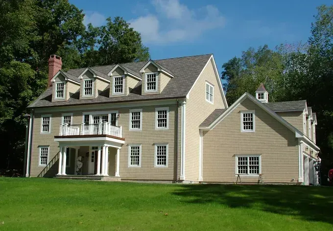 Beige two-story house with white trim, a porch, and several dormers, set on green lawn with trees in the background.