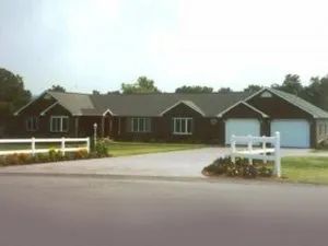Brown ranch-style house with two-car garage, white fence, and long driveway on a sunny day.