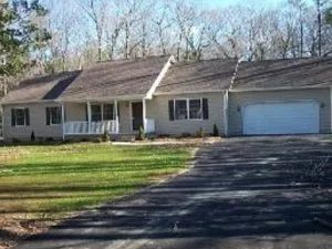 Ranch-style house with gray siding, a dark driveway, and a two-car garage. Set in a grassy yard, surrounded by trees.