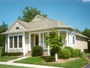 Beige house with porch, shrubs, and green lawn under a blue sky.