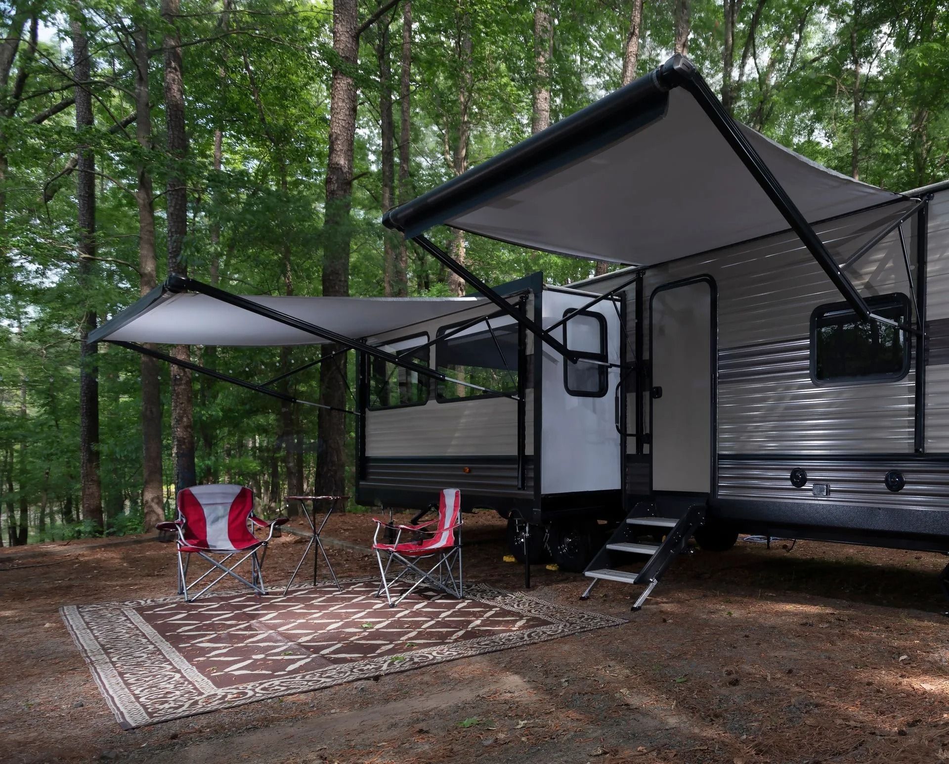Camper with awnings extended, chairs on a rug, set up in a wooded campsite.