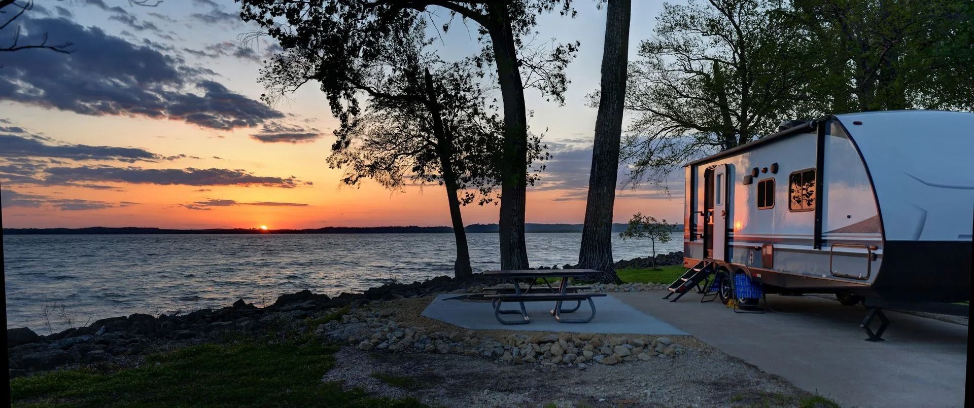 Sunset over a lake with a campsite. A trailer is parked near a picnic table and trees.
