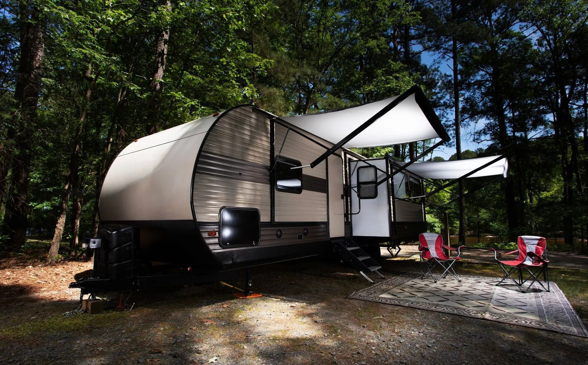RV camper with awning extended in a wooded campsite, red chairs on a rug.