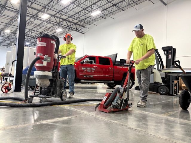 Two workers in a large warehouse grinding concrete floor. One operates a grinder, the other controls a dust collector.