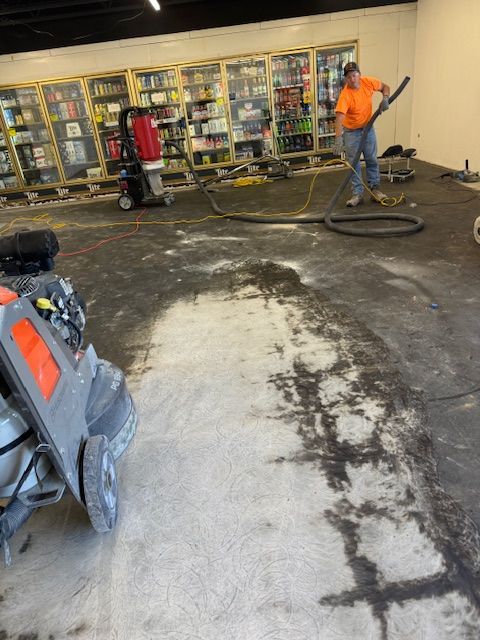 Person vacuuming a commercial floor. Machines and merchandise shelves in the background. The floor has a large wet area.