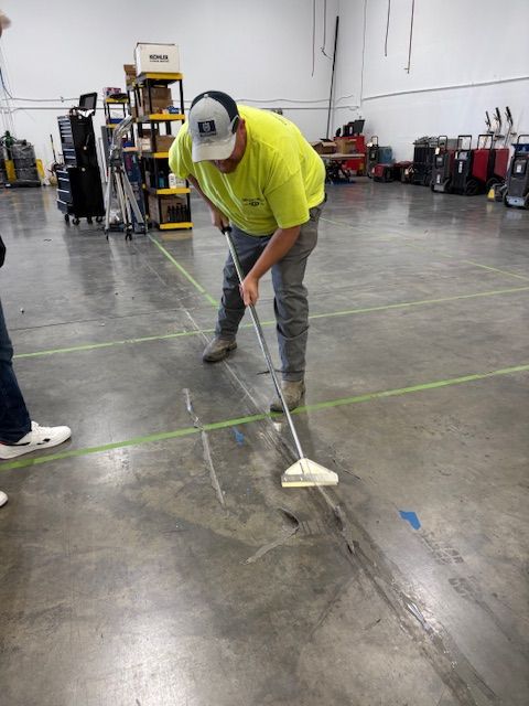 Person in a yellow shirt sweeping a concrete floor marked with tape, likely in a warehouse or workshop.