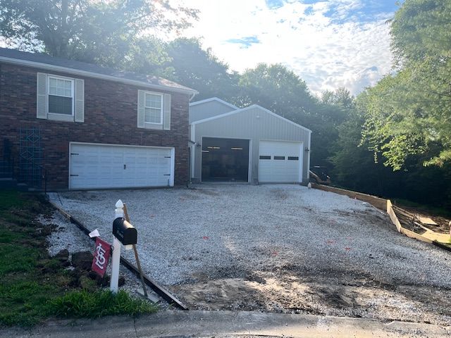 Driveway with gravel, leading to a house with a brick facade and a detached garage, set amongst trees.