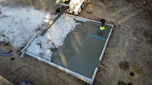 Concrete being poured from a mixer truck onto a prepared rectangular foundation. A worker levels the wet concrete.