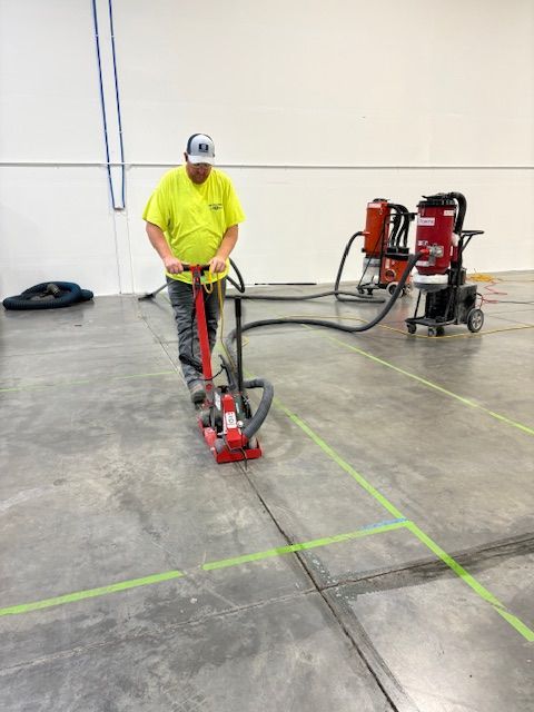 Man grinding concrete floor with machine, connected to vacuum. Inside a building, green lines on floor.