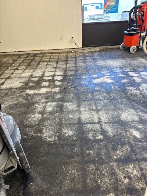 Concrete floor with a grid pattern, showing a grinding process; a vacuum and other tools are present.
