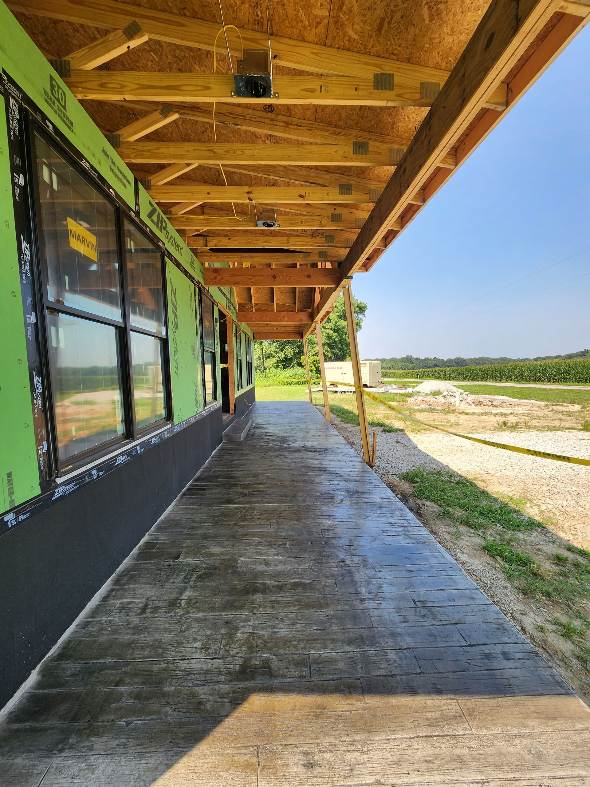 Long, covered concrete walkway under construction; windows on the left, clear sky, and rural landscape in background.