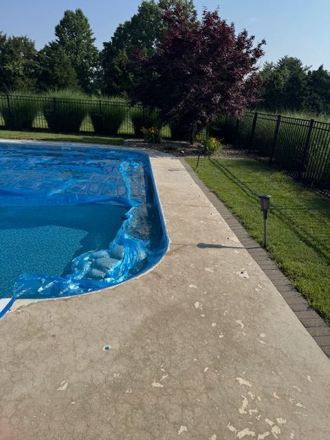 A swimming pool with blue cover, concrete deck, green grass, and trees under a clear sky.