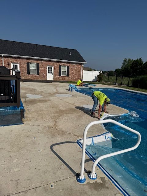 Two workers install a blue pool cover on a concrete patio next to a house under a clear sky.