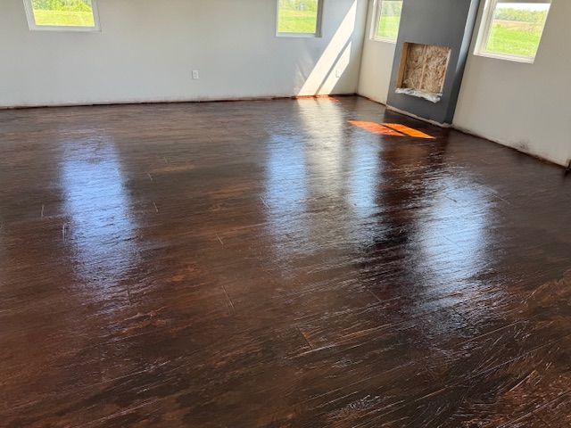 Dark stained, glossy wood floor in a room with a fireplace and windows, reflecting sunlight.