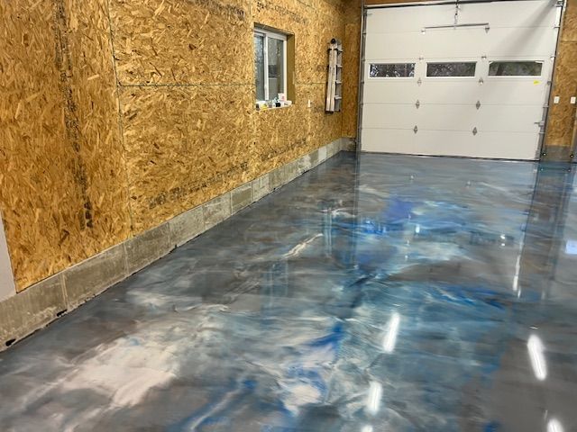 Garage with shiny epoxy floor in shades of blue and gray. Particleboard walls, concrete base, and white garage door.