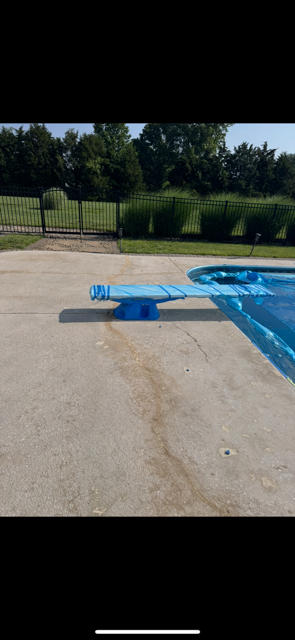 Blue diving board on concrete next to a pool. Trees and fence in the background.
