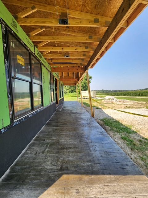 Covered concrete walkway under construction; windows on the left; fields in the background.