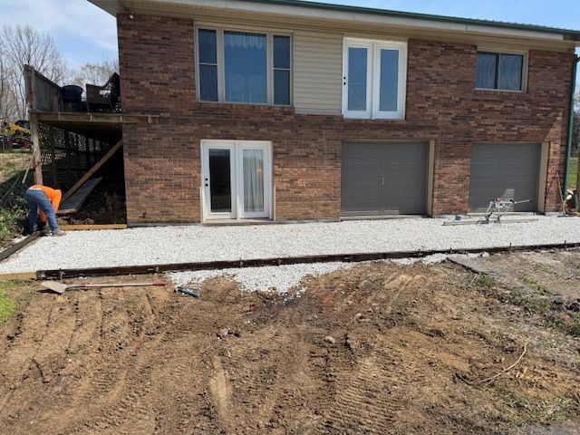Construction of a gravel patio next to a two-story brick building with garage doors. A person works on the patio.