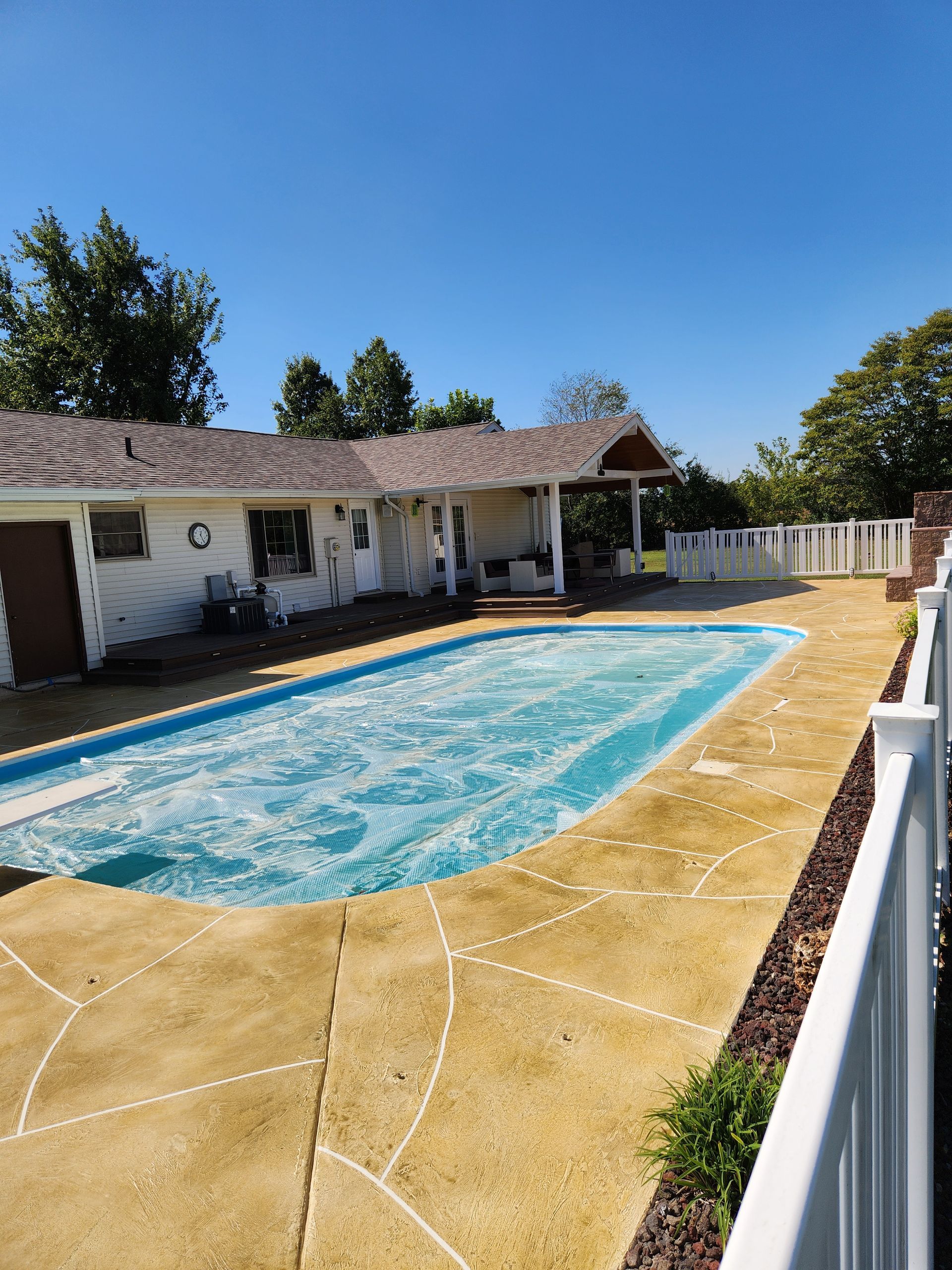 A backyard with a pool covered by a blue tarp, a white house, and a white fence on a sunny day.