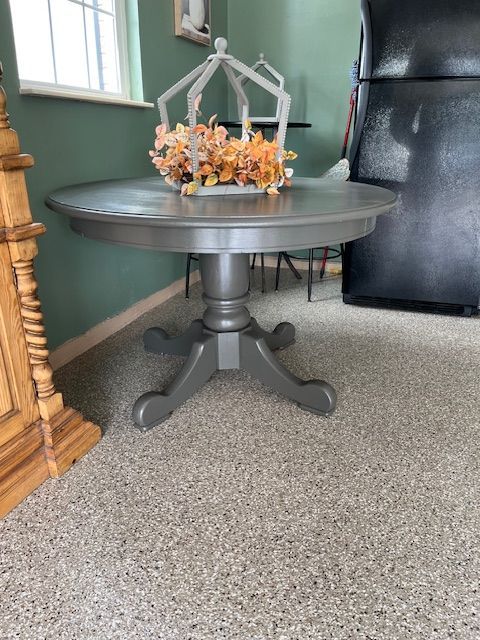 Round gray dining table with a floral centerpiece, by a window, near a refrigerator and cabinet.
