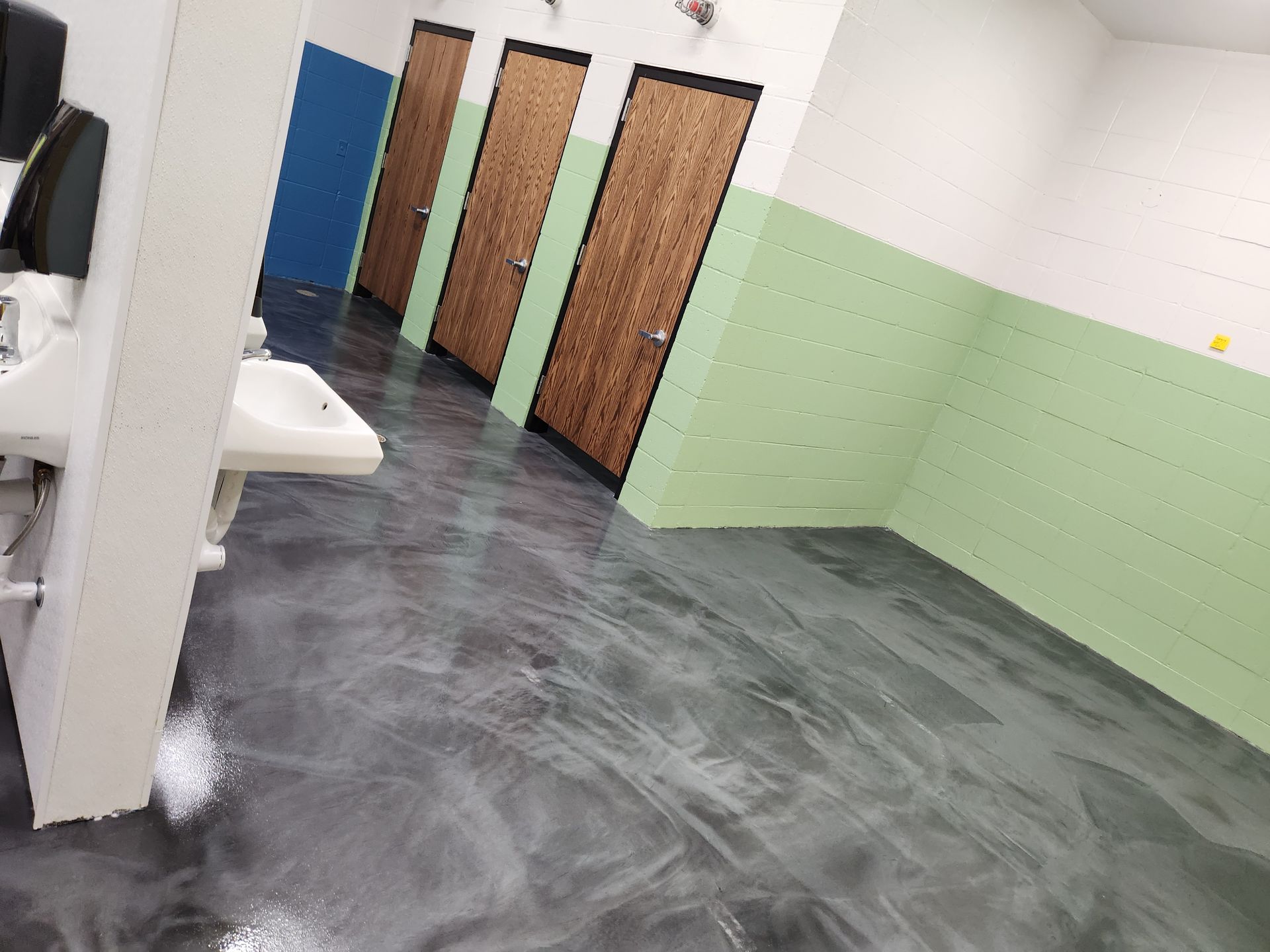 Bathroom with metallic gray floor, green and white walls, brown stall doors, and a blue door.