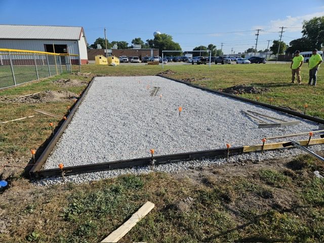 Gravel-filled rectangle with black borders, prepared for a concrete pour; two workers stand nearby.