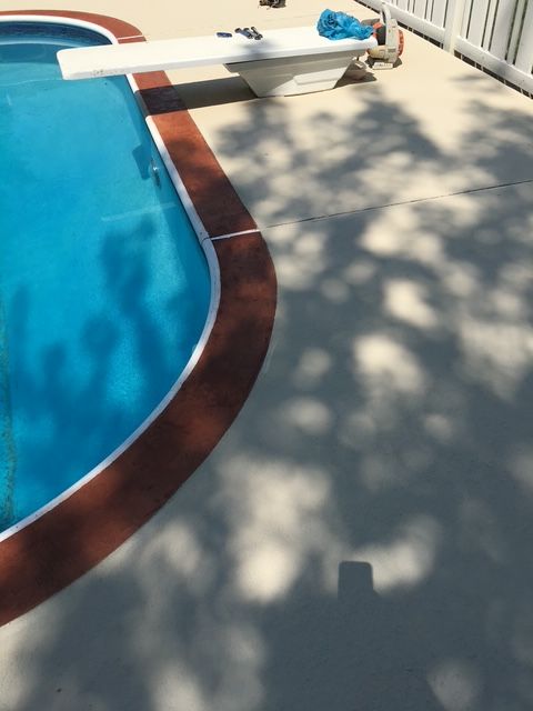 Poolside with diving board and red-brown trim bordering the blue water. Light concrete deck with tree shadows.
