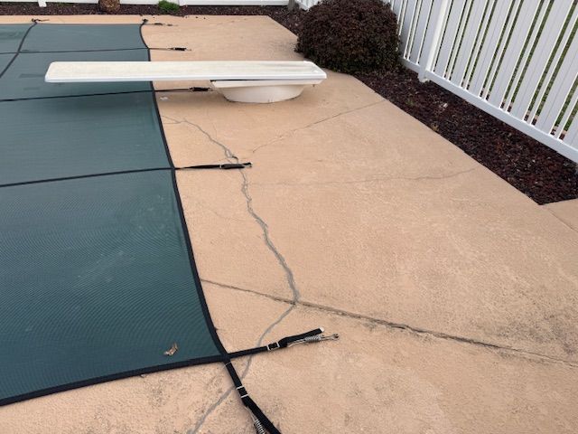 Pool cover on a concrete patio with a diving board, surrounded by a white fence and shrubbery.