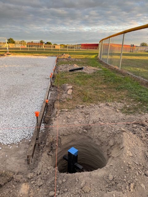 Hole dug in dirt near gravel pathway and chain-link fence on a grassy field. Construction in progress.