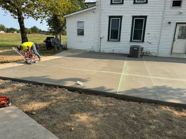 Construction worker using a saw on a newly poured concrete patio near a white house.