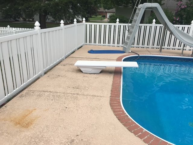 White fenced-in pool area with a diving board, water slide, and blue water. Concrete patio surrounding the pool.