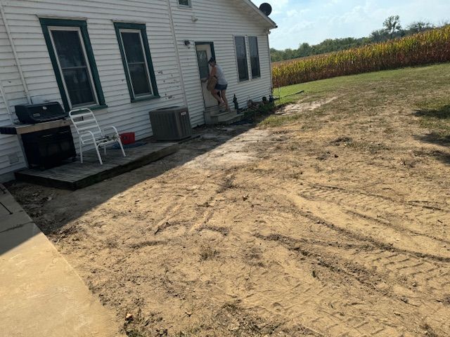 House exterior with dirt yard; person in doorway, grill, chair, air conditioner. Cornfield in background.